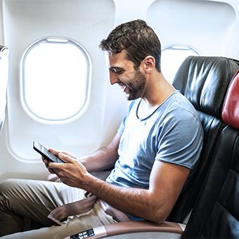 Brown haired man sitting next to a window on the plane using inflight internet on his smartphone